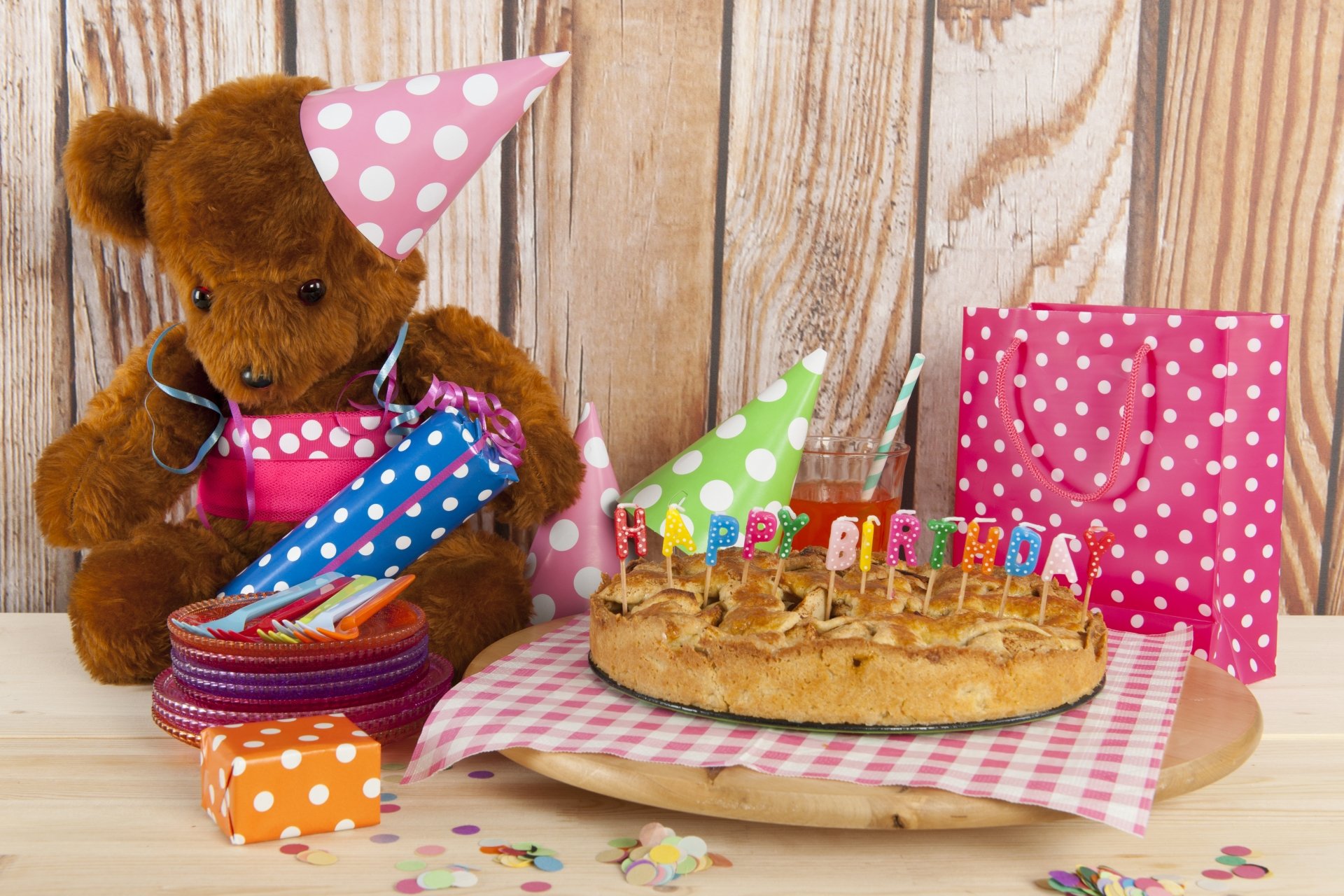 A teddy bear wearing a party hat sits beside a birthday cake with lit candles, colorful party hats, and festive gift bags on a wooden table, captured in 8K Ultra HD.