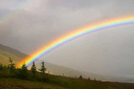 HD desktop wallpaper featuring a vibrant rainbow arching over a misty natural landscape with hills and evergreen trees.