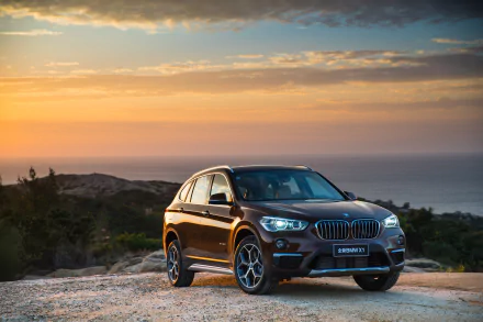 Brown BMW X1 SUV parked on a coastal overlook at sunset, ocean in the background — 4K Ultra HD PC desktop wallpaper.