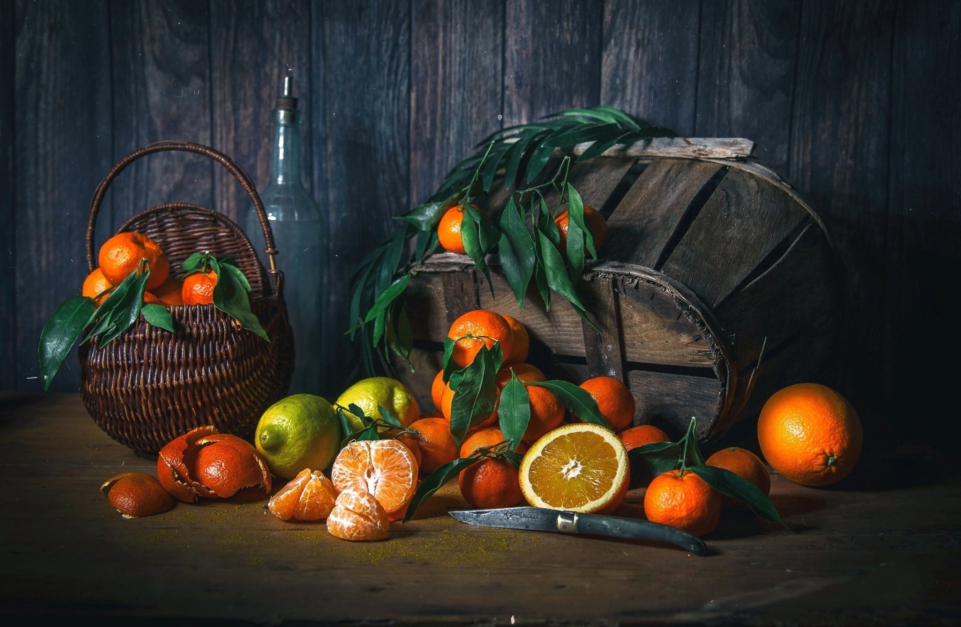 HD desktop wallpaper featuring a still life of mandarins and oranges arranged in and around a basket, with green leaves and a rustic wooden backdrop.
