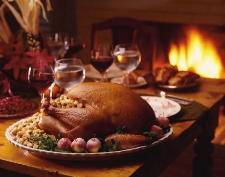 A beautifully set Thanksgiving table features a golden-brown turkey surrounded by garnishes, with elegant glassware and a warm fireplace in the background, creating a festive atmosphere.