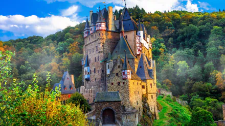 4K Ultra HD wallpaper featuring Eltz Castle, a man-made historic castle surrounded by vibrant fall foliage under a bright blue sky.