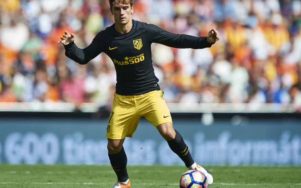 French soccer player in a dark training top and yellow shorts controls the ball on a packed stadium pitch — high-resolution 4K sports desktop wallpaper.