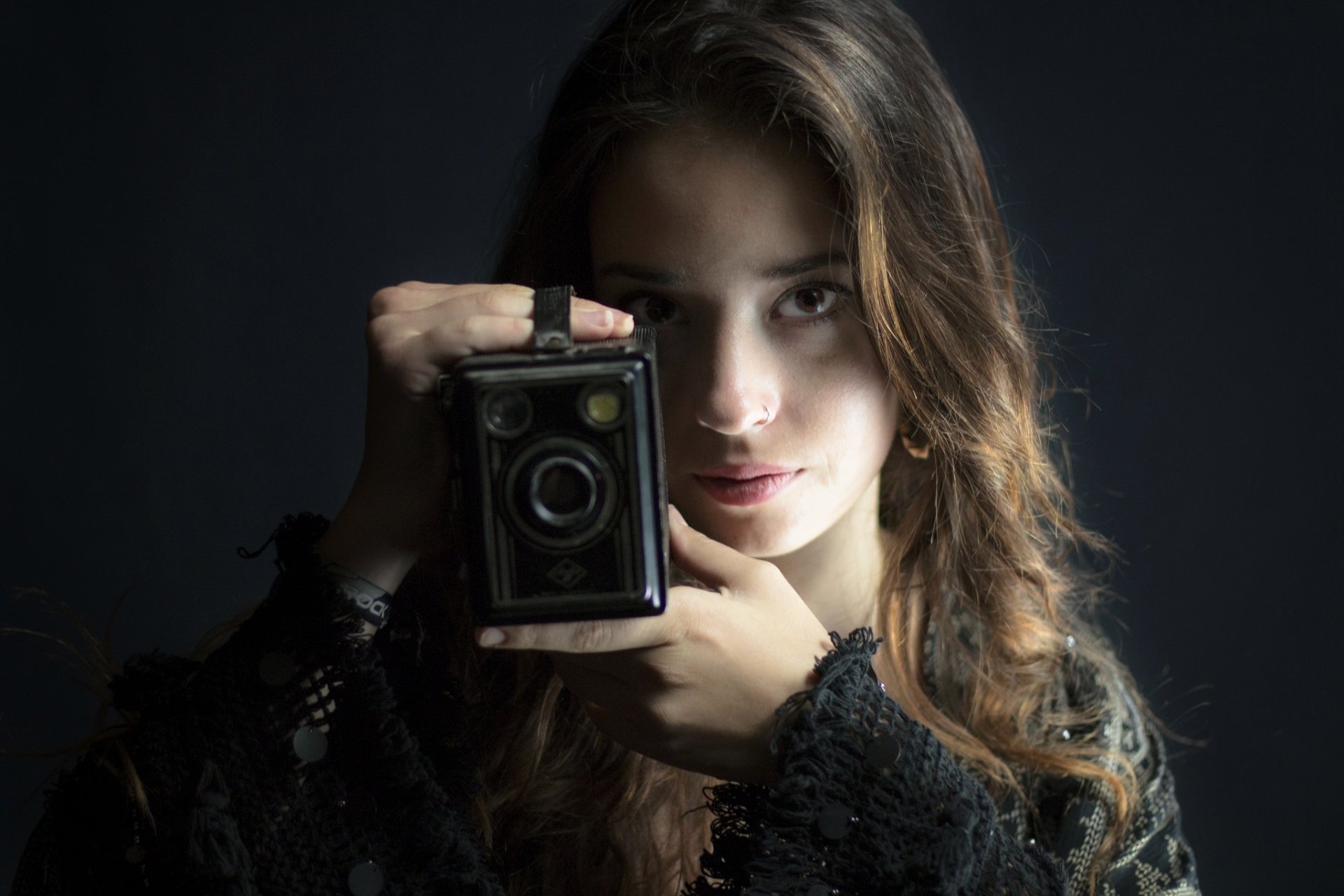 Brunette model with brown eyes holding a vintage box camera to her face, moody lighting - HD PC desktop wallpaper background.