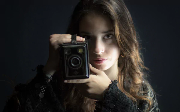 Brunette model with brown eyes holding a vintage box camera to her face, moody lighting - HD PC desktop wallpaper background.