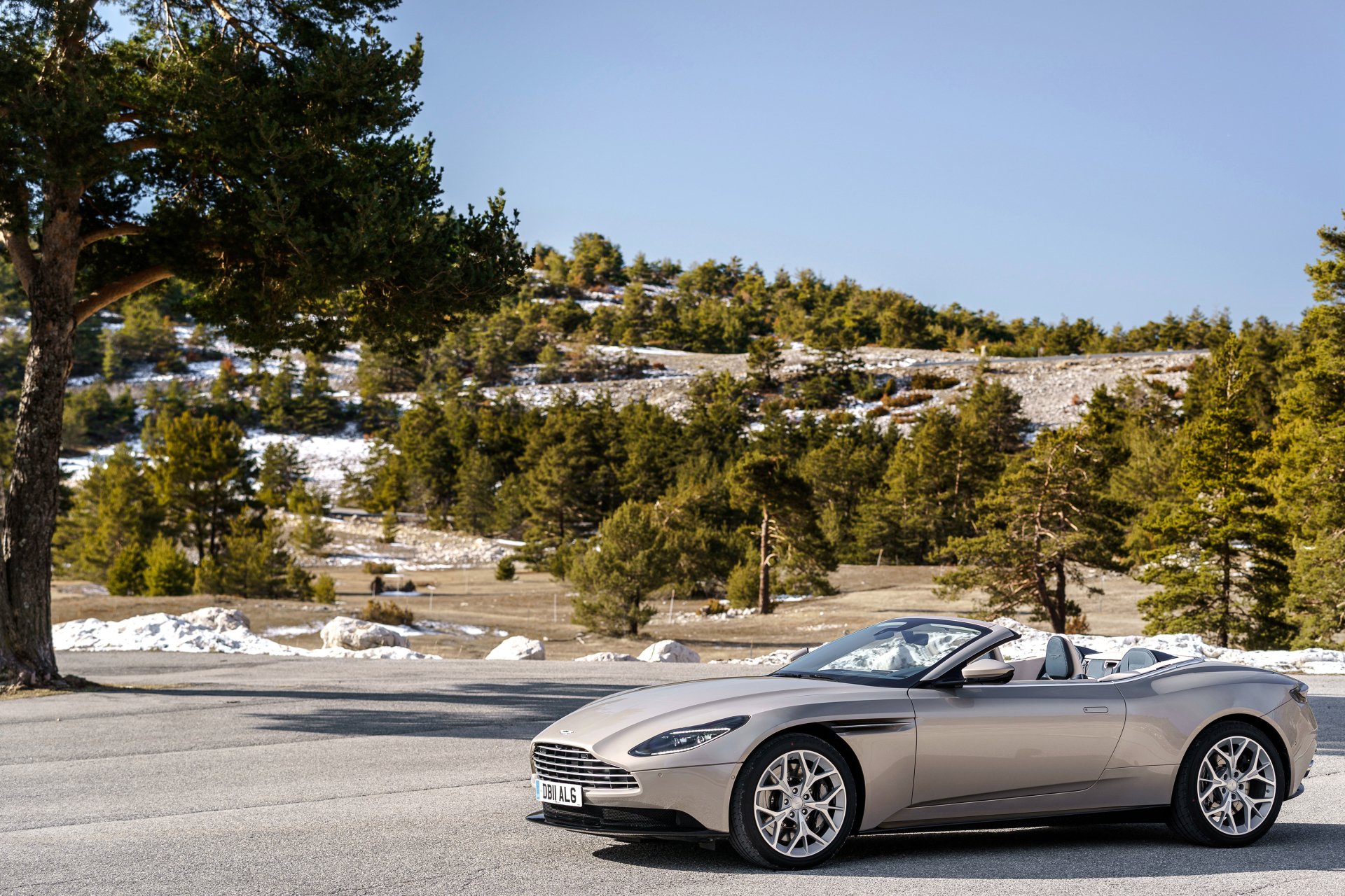 Silver Aston Martin DB11 cabriolet grand tourer on a mountain road with pines and snow, 4K Ultra HD PC desktop wallpaper.