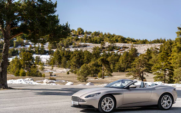Silver Aston Martin DB11 cabriolet grand tourer on a mountain road with pines and snow, 4K Ultra HD PC desktop wallpaper.