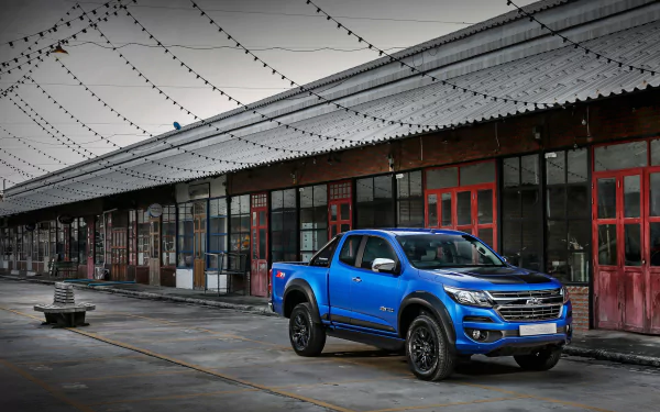 Blue Chevrolet Colorado car parked by an industrial warehouse under string lights — 4K Ultra HD PC desktop wallpaper background.
