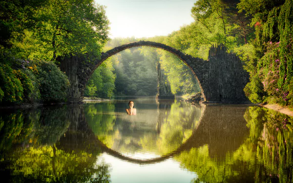 HD PC wallpaper: sunlit Devil's Bridge stone arch mirrored in a calm river, a swimmer near center, surrounded by lush trees and blue sky — manipulated photographic landscape.