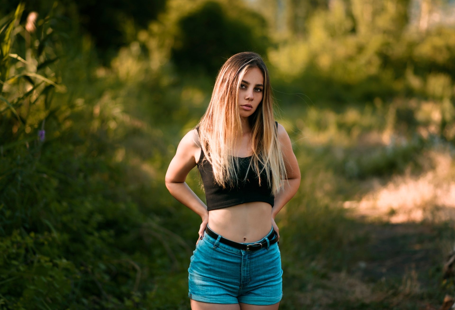 HD desktop wallpaper of a blonde woman model wearing shorts, standing outdoors with a blurred natural background and shallow depth of field.