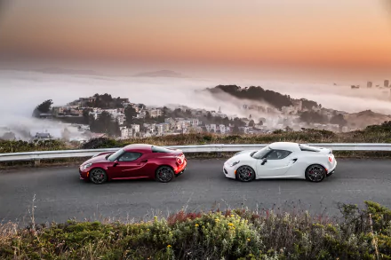 Two Alfa Romeo 4C supercars, one white and one red, parked on a mountain road with a misty cityscape at sunset, captured in an HD PC desktop wallpaper.