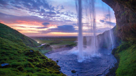 A 4K Ultra HD view of Seljalandsfoss waterfall at sunset, framed by lush green cliffs with a vibrant horizon sky over expansive nature.
