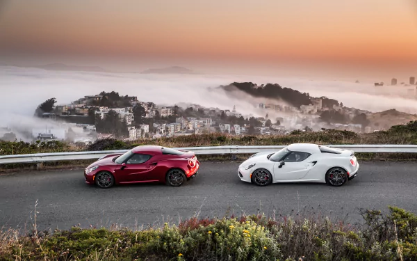 Two Alfa Romeo 4C supercars, one white and one red, parked on a mountain road with a misty cityscape at sunset, captured in an HD PC desktop wallpaper.