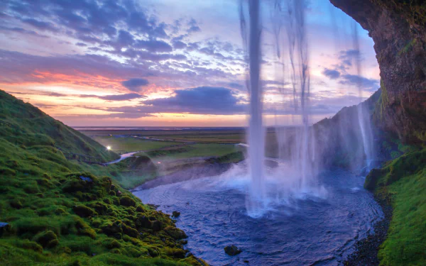 A 4K Ultra HD view of Seljalandsfoss waterfall at sunset, framed by lush green cliffs with a vibrant horizon sky over expansive nature.