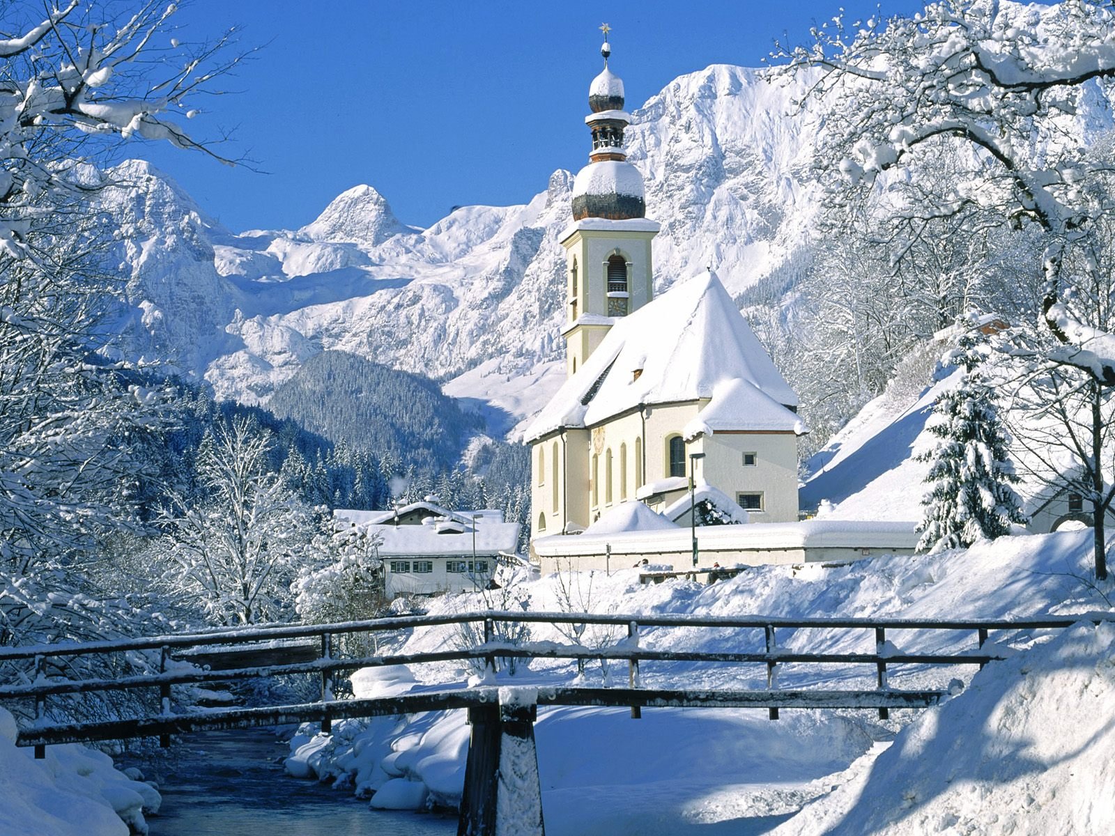 A winter scene featuring a snow-covered church beside a bridge, nestled among majestic mountains and frosted trees, creating a serene natural landscape.