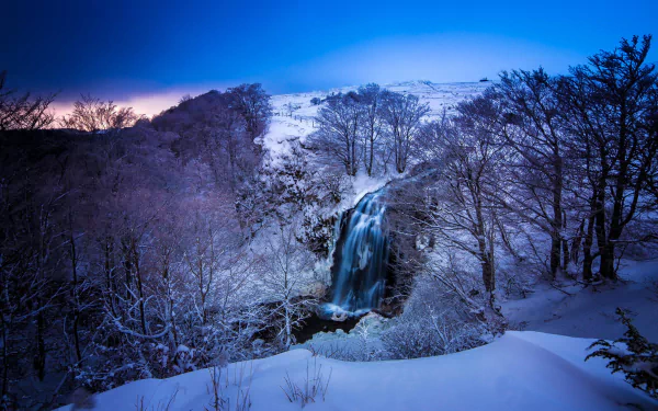 HD winter wallpaper featuring a serene forest landscape with a waterfall cascading amidst snow-covered trees. The peaceful scene captures the essence of nature in the winter season.