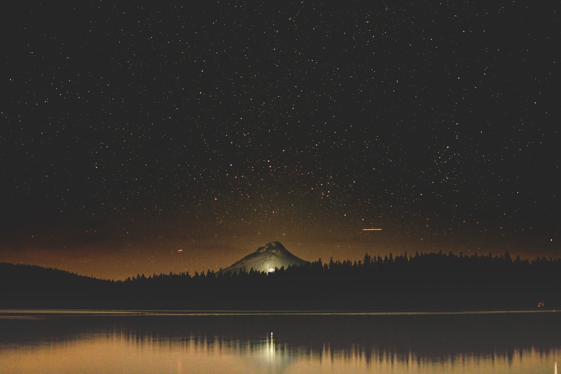 8K Ultra HD wallpaper showing a tranquil lake under a starry night sky with a softly lit hill and natural silhouettes in the background.