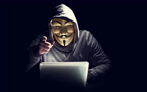 4K Ultra HD desktop wallpaper showing an anonymous figure in a hoodie and Guy Fawkes mask working on a laptop, symbolizing technology and online anonymity.