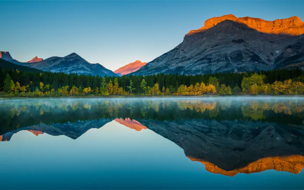 HD desktop wallpaper featuring a serene nature scene with a mountain range, lush forest, and their reflections on a calm lake. The early morning sunlight casts a golden glow on some peaks.