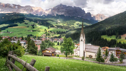Scenic view of a man-made village in Alta Badia, Dolomites, Italy, featuring a church steeple, green meadows, and rugged mountain peaks under a cloudy sky.