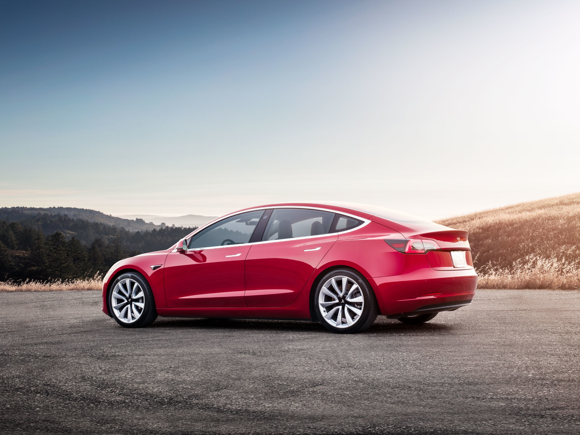 A red Tesla Model 3 parked outdoor with a scenic backdrop, serves as a sleek HD wallpaper for Tesla Motors enthusiasts.
