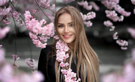 A blonde woman with long hair and blue eyes smiles amidst blooming pink flowers, creating a serene and vibrant HD desktop wallpaper background.