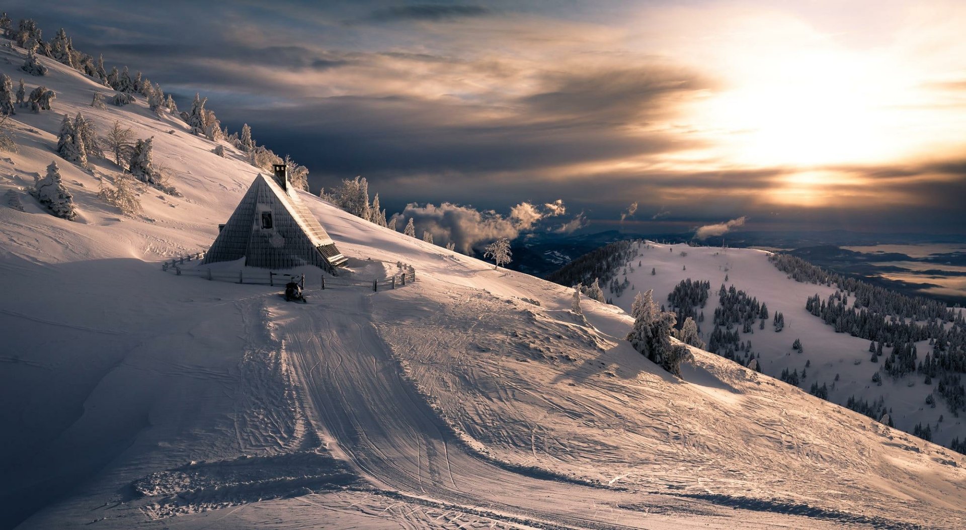 Snow-covered mountain landscape at sunset with a pine tree, a man-made cabin, and a serene winter atmosphere, captured in HD for a PC desktop wallpaper.