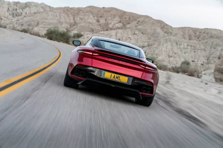 HD desktop wallpaper featuring a red Aston Martin DBS Superleggera supercar driving on a winding road through a rocky desert landscape.