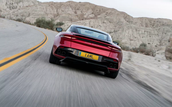 HD desktop wallpaper featuring a red Aston Martin DBS Superleggera supercar driving on a winding road through a rocky desert landscape.
