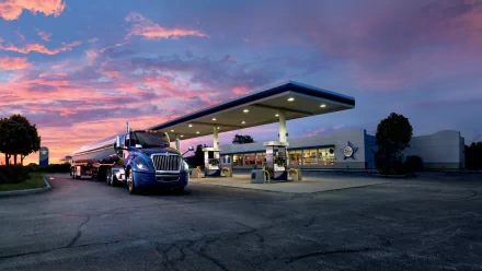 2K Quad HD PC desktop wallpaper: blue truck vehicle refueling at a roadside gas station beneath a vivid sunset sky.