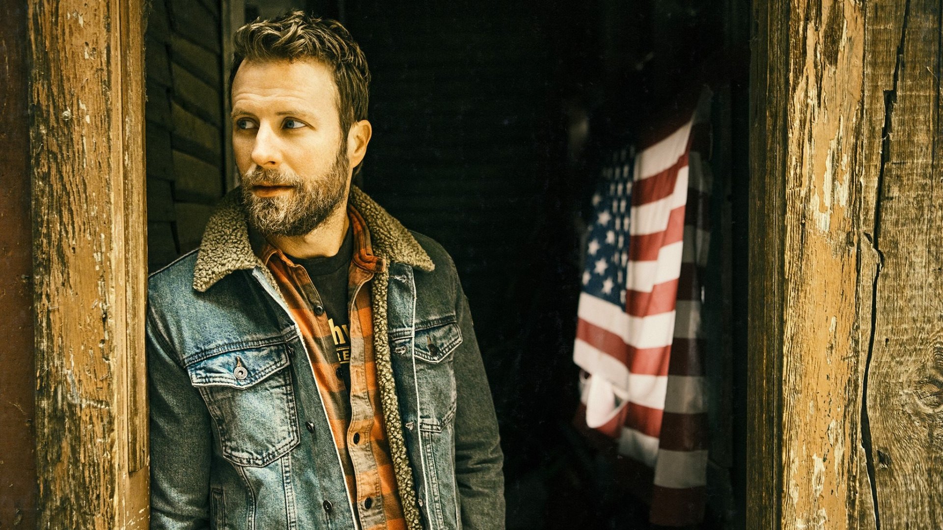 HD desktop wallpaper featuring Dierks Bentley in a denim jacket, leaning against a rustic doorway with an American flag in the background.