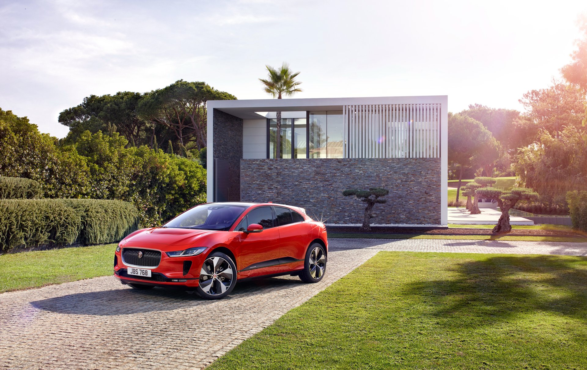 Red Jaguar I‑Pace SUV on a sunlit driveway in front of a modern house, with lawn and trees. 4K Ultra HD PC desktop wallpaper/background of a Jaguar vehicle.