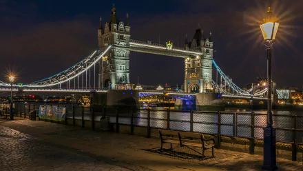 Night view of London’s Tower Bridge illuminated with lights, featuring a lamp post and empty bench by the riverside, captured in 4K Ultra HD quality.