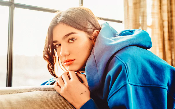 HD desktop wallpaper featuring English singer Dua Lipa with brunette hair and brown eyes, wearing a blue hoodie in a softly lit indoor setting.