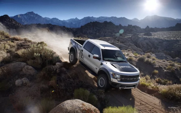 HD PC desktop wallpaper featuring a Ford Raptor vehicle driving off-road through a rugged mountainous landscape under a bright sun.
