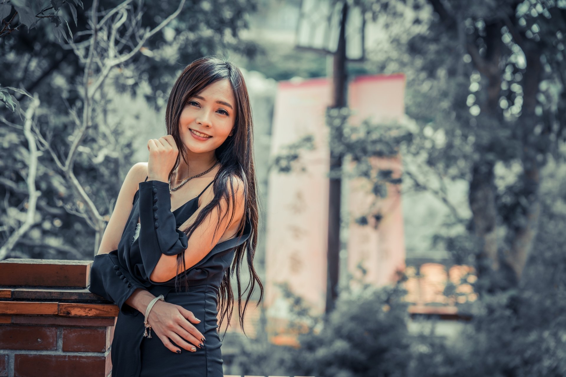 A smiling brunette Asian woman with long hair leans against a brick wall in an outdoor setting, with a softly blurred background. This HD desktop wallpaper exudes a natural and serene beauty.