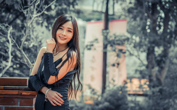 A smiling brunette Asian woman with long hair leans against a brick wall in an outdoor setting, with a softly blurred background. This HD desktop wallpaper exudes a natural and serene beauty.