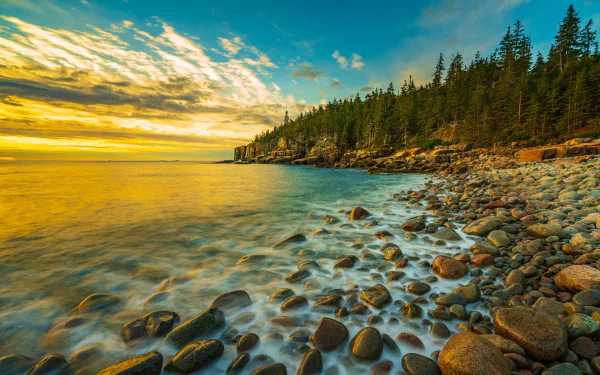 HD PC desktop wallpaper of Maine's rocky coastline at sunset, featuring ocean waves, a forested shoreline, and a vibrant sky over a natural beach setting.