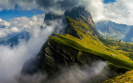 HD wallpaper of Italy’s Dolomites mountains partly covered by clouds, showcasing vibrant green slopes and dramatic peaks surrounded by mist and blue sky.