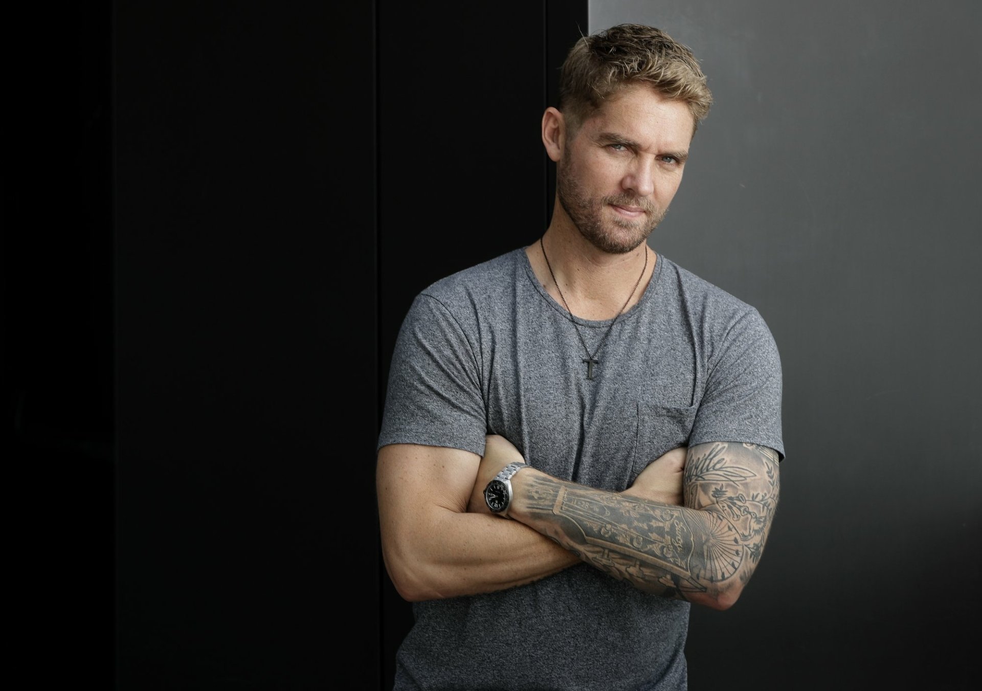 HD wallpaper featuring a confident man with crossed arms and tattoos, set against a dark background.
