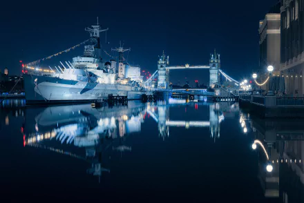 A night view of London featuring HMS Belfast (C35) and Tower Bridge, beautifully reflected in the water. The image serves as an HD desktop wallpaper and background.