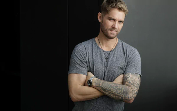 HD wallpaper featuring a confident man with crossed arms and tattoos, set against a dark background.